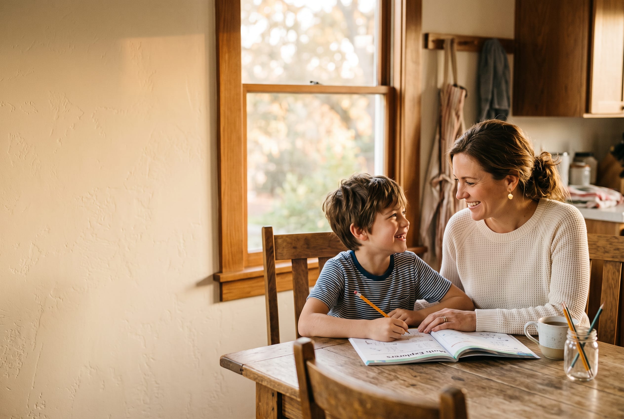 A mother and her young son sit together at a warm wood kitchen table in a Central Valley home, sharing a relaxed smile while working through a workbook in golden window light